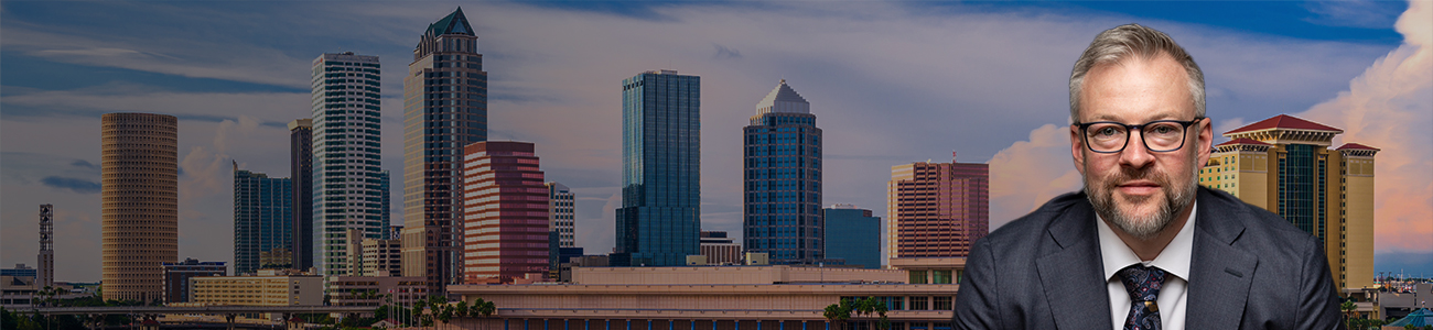 Professional portrait of Jason M. Mayberry, in a suit and tie, set against a panoramic background of Tampa's city skyline at dusk.